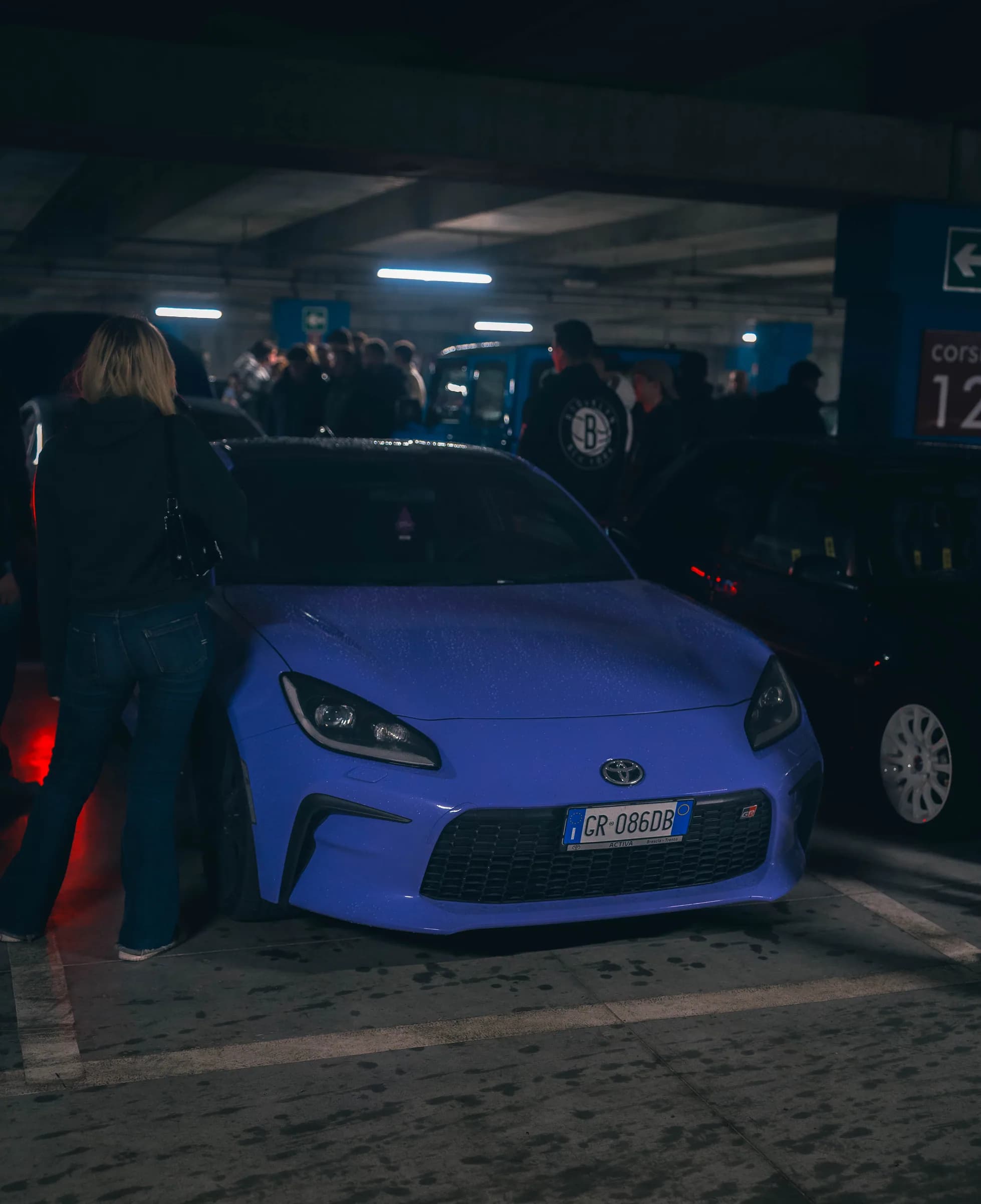 Una Toyota GR86 blu in un parcheggio coperto durante un raduno notturno, circondata da appassionati.