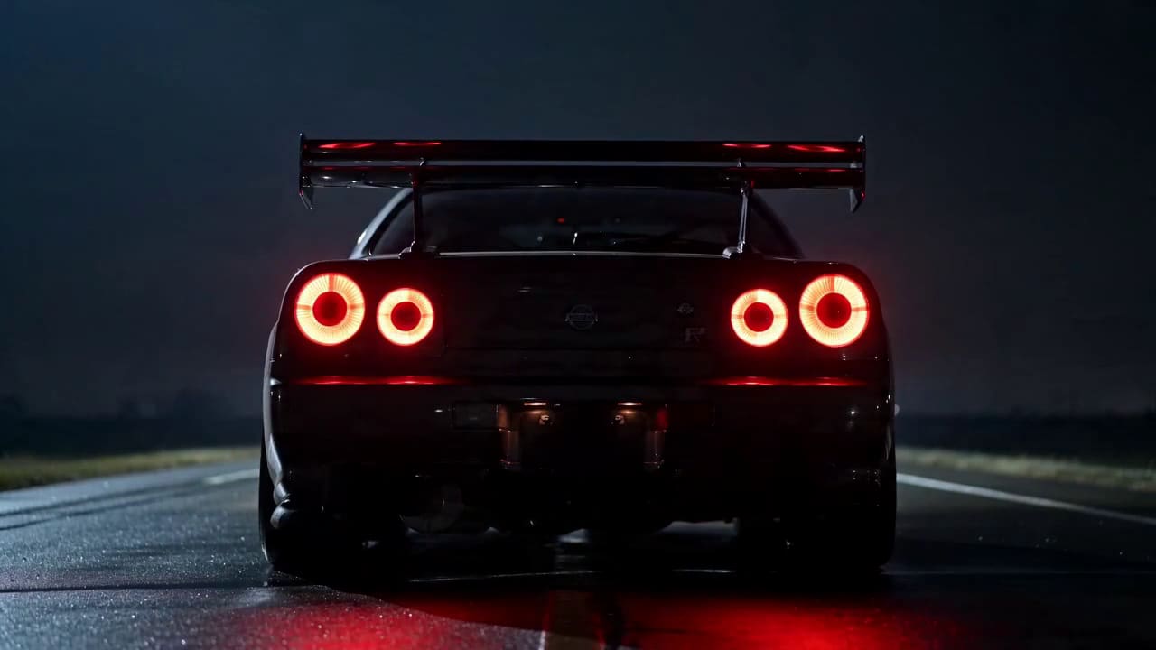 Una Nissan Skyline GT-R R34 vista da dietro su una pista bagnata di notte, con i quattro fari posteriori accesi.